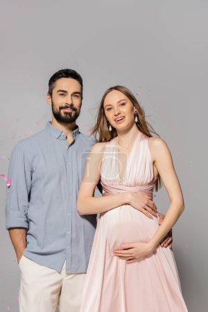 Positive man hugging elegant and pregnant wife in dress and looking at camera while standing under confetti during baby shower party on grey background, expecting parents concept 