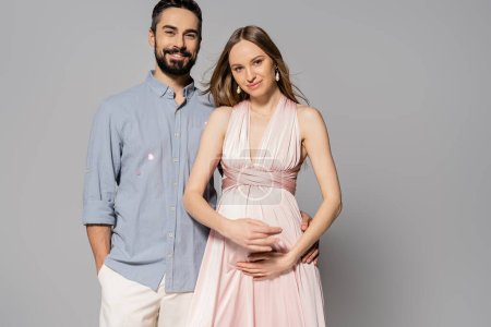 Stilvoller und fröhlicher Mann umarmt elegante und schwangere Frau, während er in die Kamera blickt und auf grauem Hintergrund steht, erwartungsvolle Eltern, Neuanfang, Elternschaft 