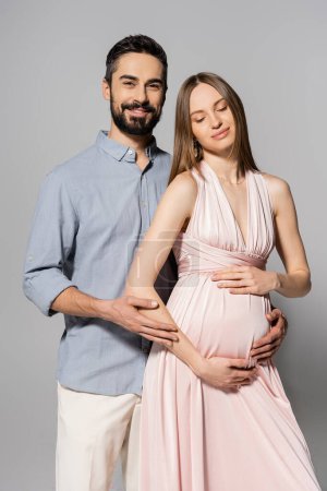 Positiver Mann umarmt elegante und schwangere Frau im rosafarbenen Kleid und blickt in die Kamera, während er vor grauem Hintergrund zusammen steht, werdende Eltern, Neuanfang 