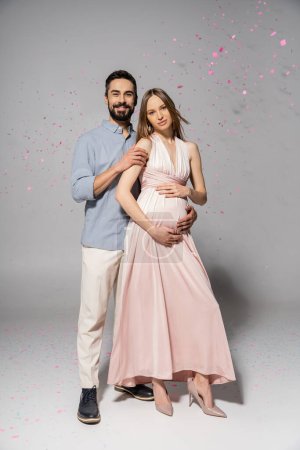 Full length of positive and elegant man hugging pregnant wife in dress while standing under confetti during baby shower party on grey background, expecting parents concept 