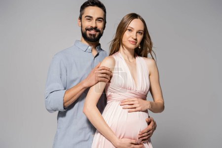 Portrait of positive and bearded man hugging elegant pregnant wife in pink dress while standing together isolated on grey, expecting parents concept, new beginnings, excitement 
