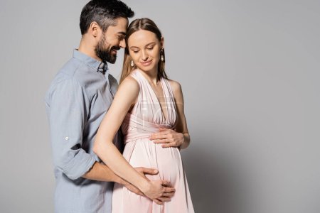Glücklicher und bärtiger Mann umarmt schwangere Frau in elegantem Kleid, während er gemeinsam auf grauem Hintergrund steht und Eltern, Ehemann und Ehefrau erwartet 