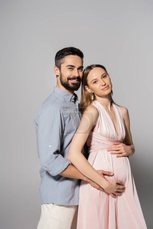 Lächelnder bärtiger Mann umarmt elegante Schwangere in rosa Kleid und blickt gemeinsam in die Kamera auf grauem Hintergrund, werdende Eltern, Ehemann und Ehefrau 