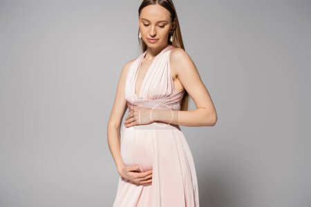 Elegante und hellhaarige werdende Mutter im rosa Kleid, die den Bauch berührt und auf grauem Hintergrund nach unten schaut, Konzept der Umstandsmode, modische Schwangerschaftskleidung 