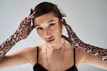spring fashion, portrait of appealing asian woman with bold makeup, brunette hair and trendy hairstyle posing in animal print gloves, silver accessories and black strap dress on grey background