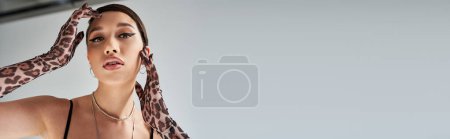 Foto de Moda de primavera, retrato de atractiva mujer asiática con maquillaje audaz, en collares de plata, pendientes y guantes de impresión animal mirando a la cámara sobre fondo gris, bandera - Imagen libre de derechos