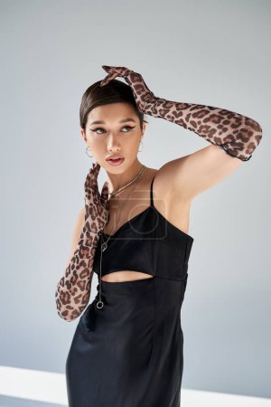 appealing asian woman with brunette hair and bold makeup posing in black strap dress and animal print gloves on grey background, spring style, modern fashion photography