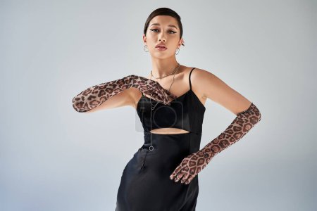 Photo for Young and graceful asian woman with brunette hair and bold makeup posing in black strap dress, animal print gloves and silver necklaces on grey background, spring fashion photography - Royalty Free Image