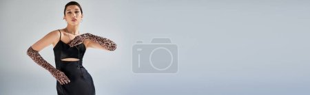Foto de Mujer asiática joven y encantadora con maquillaje audaz y cabello moreno de pie en pose elegante sobre fondo gris, moda de primavera, vestido de correa negra, guantes de impresión animal, generación z, bandera - Imagen libre de derechos