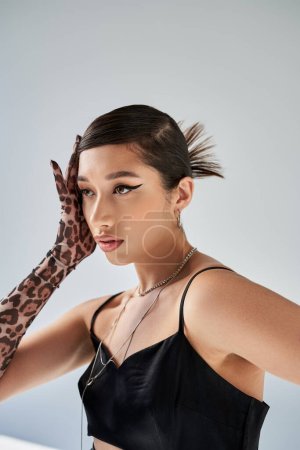 sensual asian fashion model with bold makeup and trendy hairstyle, wearing black strap dress, animal print glove and silver accessories, holding hand near face on grey background, stylish spring