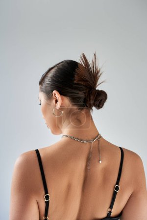 Frühlingsmode, sinnliche Frau mit brünetten Haaren und trendiger Frisur, in silbernen Halsketten und Ohrringen, posiert im schwarzen Trägerkleid auf grauem Hintergrund, Generation Z, Rückseite