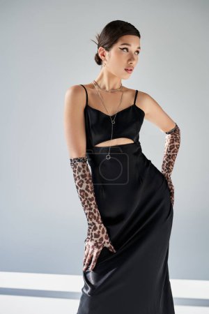 spring style, fashion shoot, young and elegant asian woman with trendy hairstyle, in black strap dress and animal pring gloves posing with hand on hip on grey background, generation z