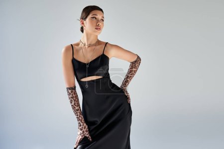 spring style, expressive asian woman with brunette hair and bold makeup standing with hand on hip in black elegant strap dress, animal print gloves and silver necklaces on grey background