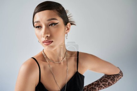 young and attractive asian woman with brunette hair, bold makeup and trendy hairstyle posing in black strap dress, silver necklaces and earrings on grey background, spring fashion photography