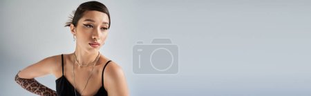 retrato de mujer asiática atractiva con cabello moreno y maquillaje audaz en vestido de correa negra, collares de plata y guante de impresión animal sobre fondo gris, estilo primavera, sesión de moda, bandera