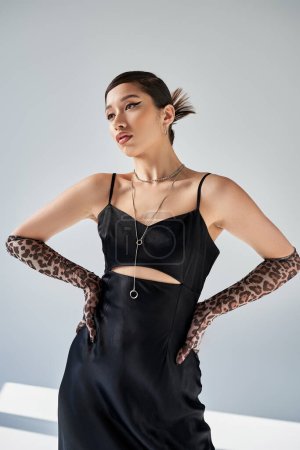 young and expressive asian woman with bold makeup posing in trendy spring outfit, black strap dress, animal print gloves, silver necklaces, fashion shoot on grey background