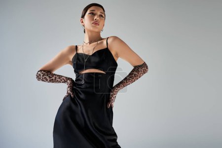 low angle view of brunette asian woman with bold makeup posing with hands on hips on grey background, black elegant strap dress, animal print gloves, silver accessories, spring fashion photography