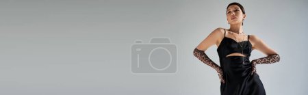 Foto de Vista de ángulo bajo de modelo de moda asiática con cabello moreno y maquillaje audaz posando con las manos en las caderas sobre fondo gris, estilo primavera, vestido de correa negra, guantes de impresión animal, pancarta - Imagen libre de derechos