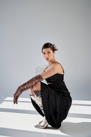 Photo for Full length of young asian woman in elegant spring outfit posing on haunches on grey background with lighting and shadows, black strap dress, animal print gloves, silver boots, generation z - Royalty Free Image