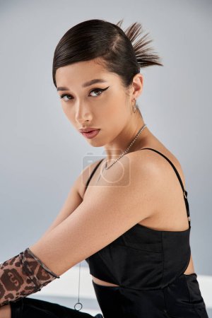 spring style asian woman with bold makeup, brunette hair and expressive gaze looking at camera on grey background, black strap dress, silver necklaces, animal print glove, fashion shoot