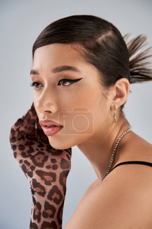 portrait of mesmerizing asian woman with brunette hair, bold makeup, trendy hairstyle, silver necklaces and animal print glove posing with hand near face on grey background, spring fashion