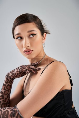 portrait of fashionable asian woman with trendy hairstyle and bold makeup posing in black dress, animal print gloves and silver necklaces while looking away on grey background, stylish spring
