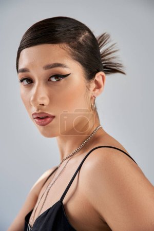 Téléchargez les photos : Printemps mode shoot, portrait de femme asiatique envoûtante en robe à bretelles noires, avec cheveux bruns, maquillage audacieux, coiffure tendance et accessoires argentés regardant la caméra sur fond gris - en image libre de droit