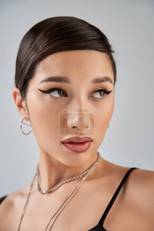portrait of appealing and young asian woman in silver accessories, with brunette hair, bold makeup and trendy hairstyle looking away on grey background, stylish spring, fashion shoot