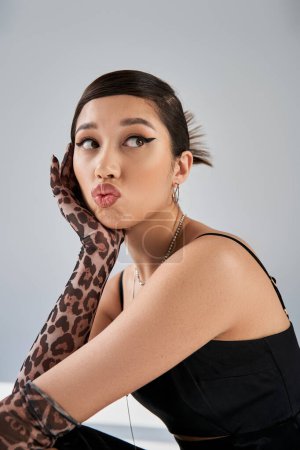 portrait de femme asiatique à la mode avec maquillage audacieux et cheveux bruns boudant les lèvres et regardant loin sur fond gris, mode de printemps, robe noire, gants d'impression animale, accessoires argentés