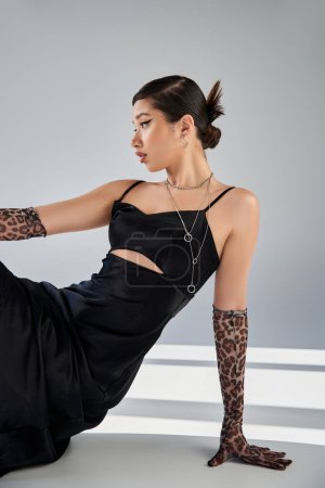 expressive asian woman with brunette hair and bold makeup, in silver necklaces, animal print gloves and black dress sitting on grey background with lighting and shadows, fashionable spring 