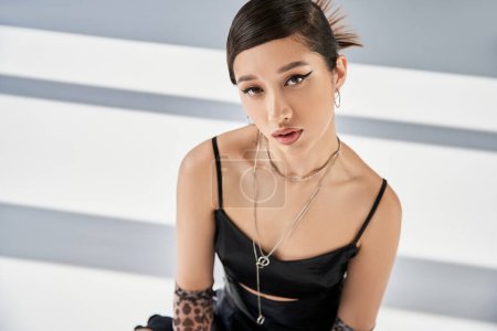 spring fashion, attractive asian woman with bold makeup, trendy hairstyle and silver accessories posing in black strap dress and looking at camera on grey background with lighting and shadows