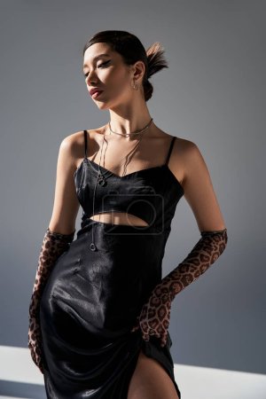 young and elegant asian woman in trendy spring outfit posing on grey background with lighting, black elegant dress, animal print gloves, silver accessories, generation z