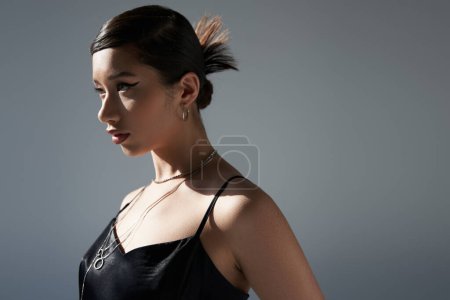 portrait of youthful asian woman with brunette hair, bold makeup and silver accessories posing in black strap dress in lighting on grey background, spring fashion concept