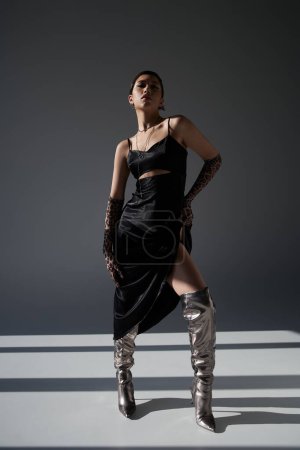full length of graceful asian woman in black strap dress, animal print gloves and silver boots standing with hand on hip on dark grey background with lighting, spring outfit, youthful style