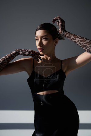trendy spring, graceful asian woman in black strap dress and animal print gloves standing in stylish pose on dark grey background with lighting, spring fashion photography