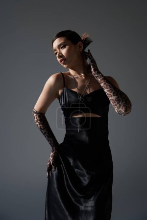 youthful fashion, stylish spring, sensual asian woman with brunette hair, in black elegant dress and animal print gloves posing with hand on hip on dark grey background