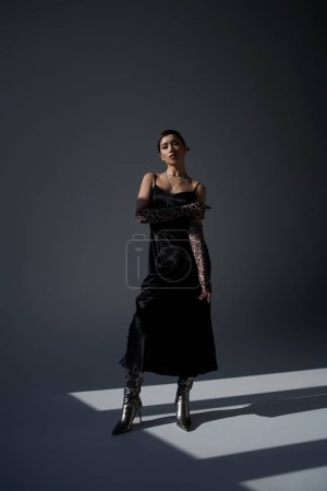 full length of young and stylish asian woman with brunette hair, in black strap dress, animal print gloves and silver boots standing on dark grey background with lighting, spring fashion photography