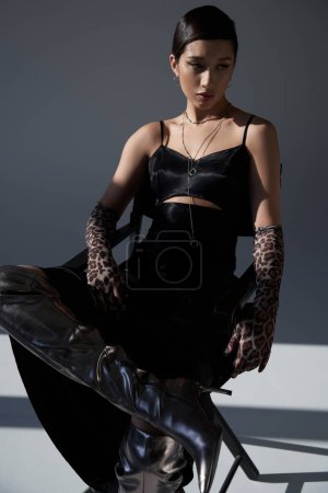 youthful and elegant asian woman in black strap dress, silver necklaces, animal print gloves and silver boots posing on chair on dark grey background with lighting, spring fashion photography
