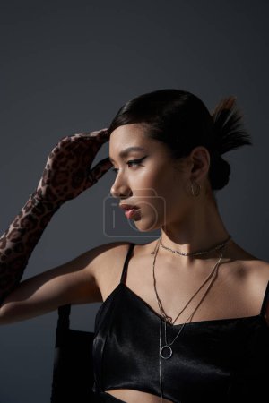 portrait of mesmerizing asian woman with brunette hair and bold makeup, in black strap dress and silver accessories sitting and holding hand near head on dark grey background, trendy spring concept