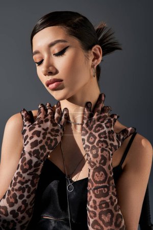 Porträt einer sinnlichen asiatischen Frau mit kühnem Make-up, im schwarzen Trägerkleid und Handschuhen mit Animal Print posiert mit geschlossenen Augen auf dunkelgrauem Hintergrund, trendiges Frühjahrskonzept, Generation z