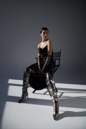 spring fashion, full length of elegant asian woman in black strap dress, animal print gloves and silver boots sitting on chair on grey background with lighting and shadows