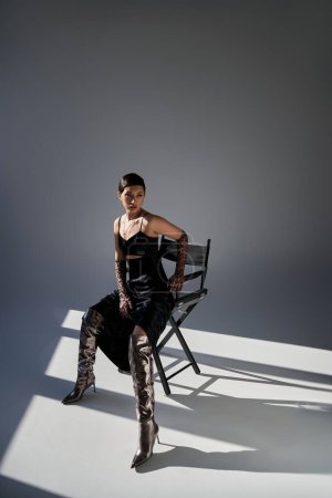 full length of young asian woman with brunette hair sitting on chair in spring outfit on grey background with lighting, black strap dress, animal print gloves, silver boots, fashion shoot