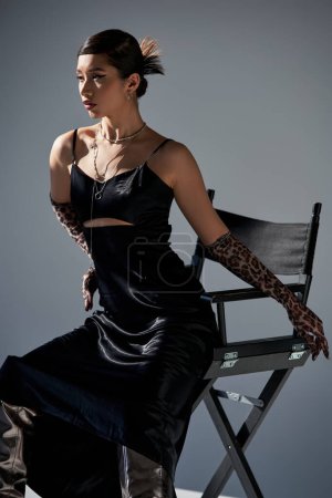 young asian woman with bold makeup and trendy hairstyle, in black elegant dress, silver necklaces and animal print gloves posing on chair on grey background with lighting, stylish spring