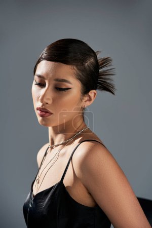 Porträt einer attraktiven Asiatin mit brünetten Haaren, fettem Make-up, trendiger Frisur, im schwarzen Trägerkleid und silberfarbenen Accessoires posiert in Beleuchtung auf grauem Hintergrund, Frühjahrsmodekonzept