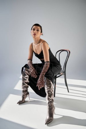 full length of sensual asian woman with expressive gaze wearing black strap dress, animal print gloves and silver boots while sitting on chair on grey background with lighting, fashionable spring