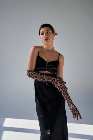 graceful asian woman with bold makeup and brunette hair standing in black strap dress and animal print gloves while looking away on grey background with lighting, stylish spring, fashion shoot