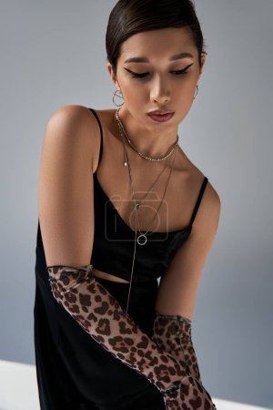 fashionable asian woman with bold makeup, brunette hair, silver necklaces and animal print gloves posing in black strap dress on grey background, stylish spring, generation z