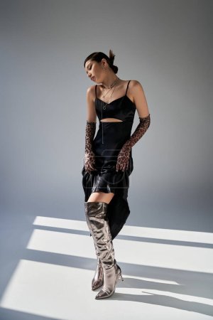 Photo for Full length of young asian woman with brunette hair standing in expressive pose in black strap dress, animal print gloves and silver boots on grey background with lighting, spring fashion concept - Royalty Free Image