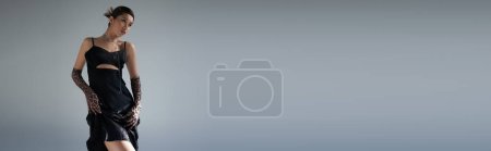 jugendliche und sinnliche asiatische Frau mit brünetten Haaren posiert in schwarzem Trägerkleid und Animal-Print-Handschuhen vor grauem Hintergrund, trendiges Frühjahrskonzept, Generation Z, Banner