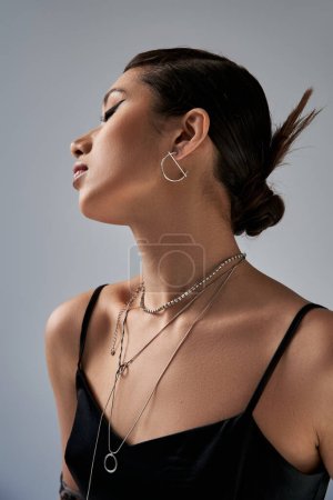 Porträt einer charmanten Asiatin mit trendiger Frisur, brünetten Haaren, fettem Make-up, silbernen Halsketten und Ohrring posiert in schwarzem Trägerkleid auf grauem Hintergrund, stylisches Frühlings-Outfit